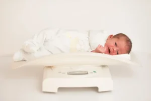 Swaddled newborn lying on a white baby scale for a weight check, representing routine monitoring of newborn weight loss and gain in the first two weeks after birth
