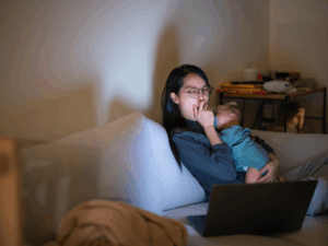Mother sitting on a couch at night holding her baby during a cluster feeding session while watching a laptop, representing the reality of frequent newborn feeding and the importance of resting and staying comfortable during the process