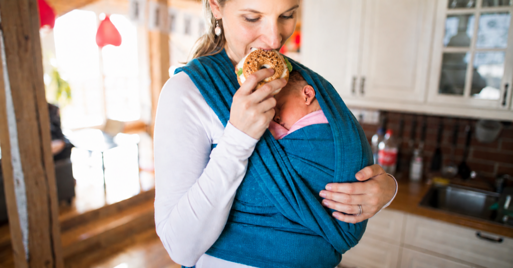 New mother wearing newborn in a blue wrap carrier while eating in the kitchen, showing the reality of postpartum life