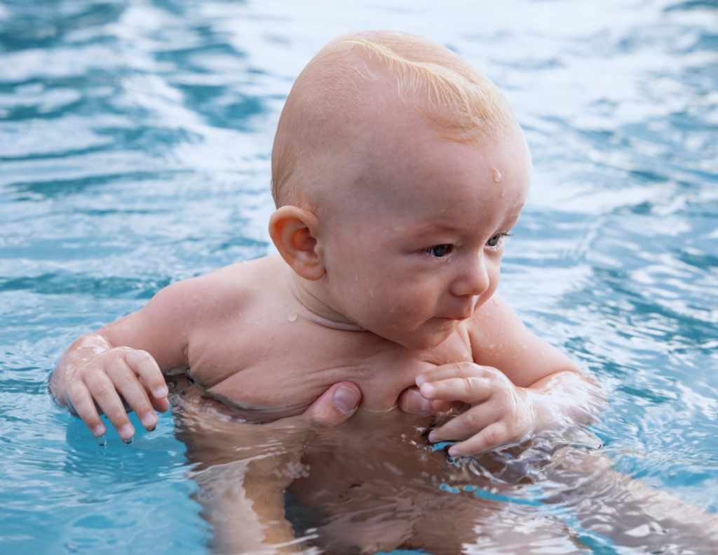 Close-up photo of a young infant supported by both of a parent's hands while floating upright in a bright blue pool, depicting safe hands-on supervision during a baby's first swimming introduction.