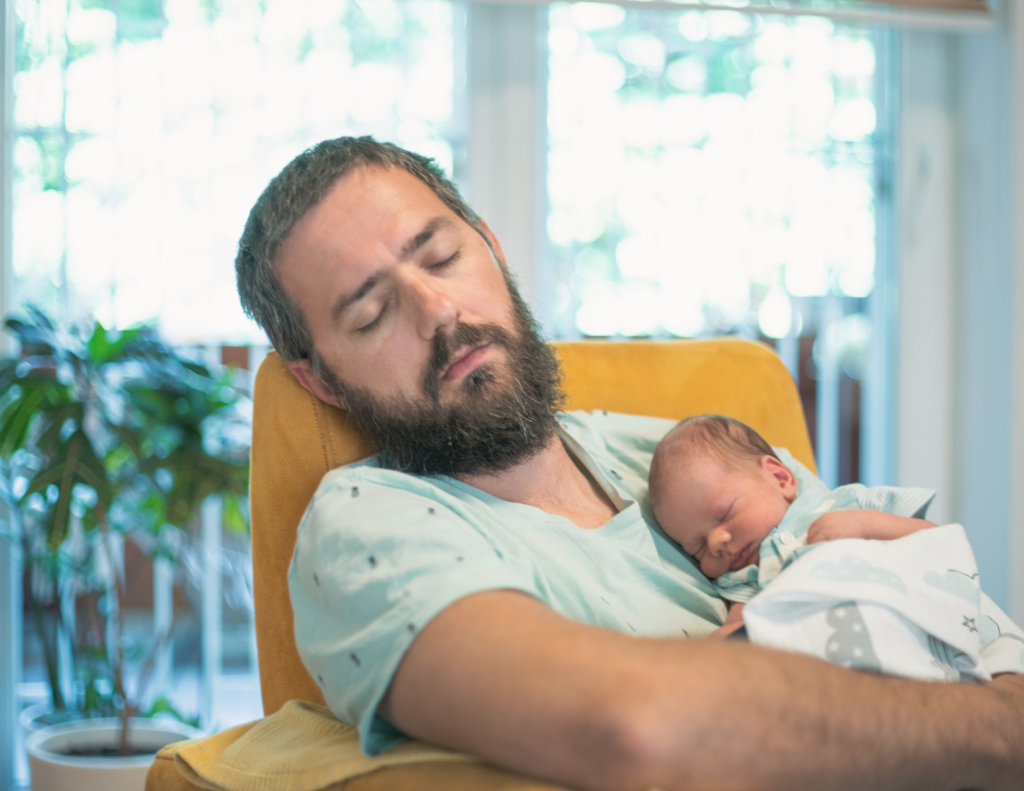 Bearded father asleep in a yellow armchair with a newborn sleeping on his chest during the day