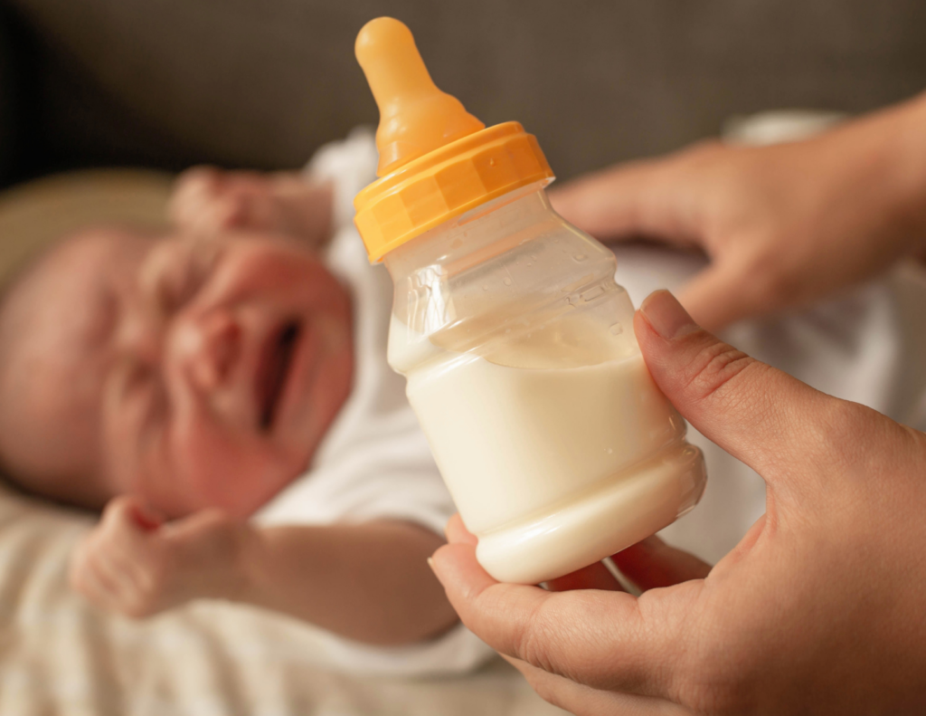 A parent holding a formula bottle in the foreground while a crying baby lies in the background