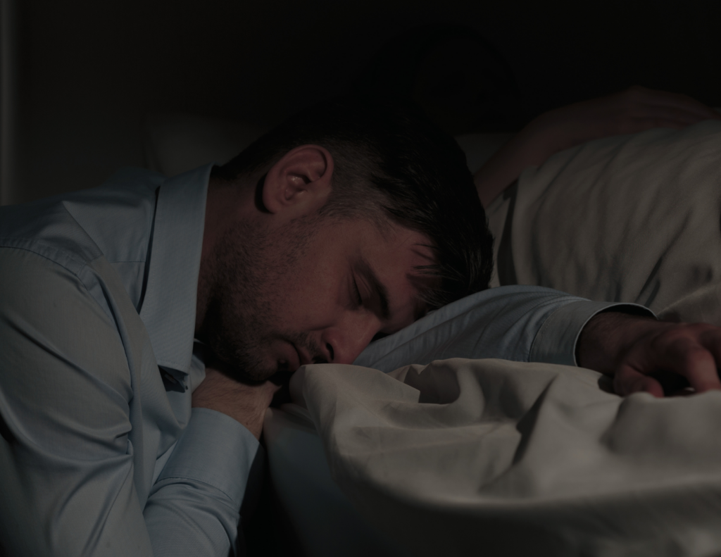 Exhausted father in a dress shirt sleeping face-down on a pillow in a dark room, still fully clothed