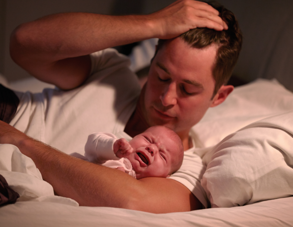 Exhausted father lying in bed at night with his hand on his head while holding a crying newborn on his chest