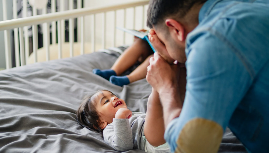 A father leaning over his smiling newborn during a playful peekaboo game. Peekaboo helps newborns and young infants develop object permanence, social bonding, and early cognitive skills. Simple face-to-face play like this supports healthy brain development in the first months of life.