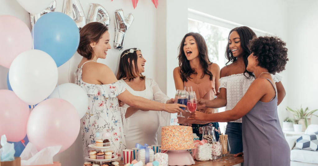 Group of diverse women laughing and toasting at a baby shower decorated with pink and blue balloons, representing the village of support new parents need in the postpartum weeks
