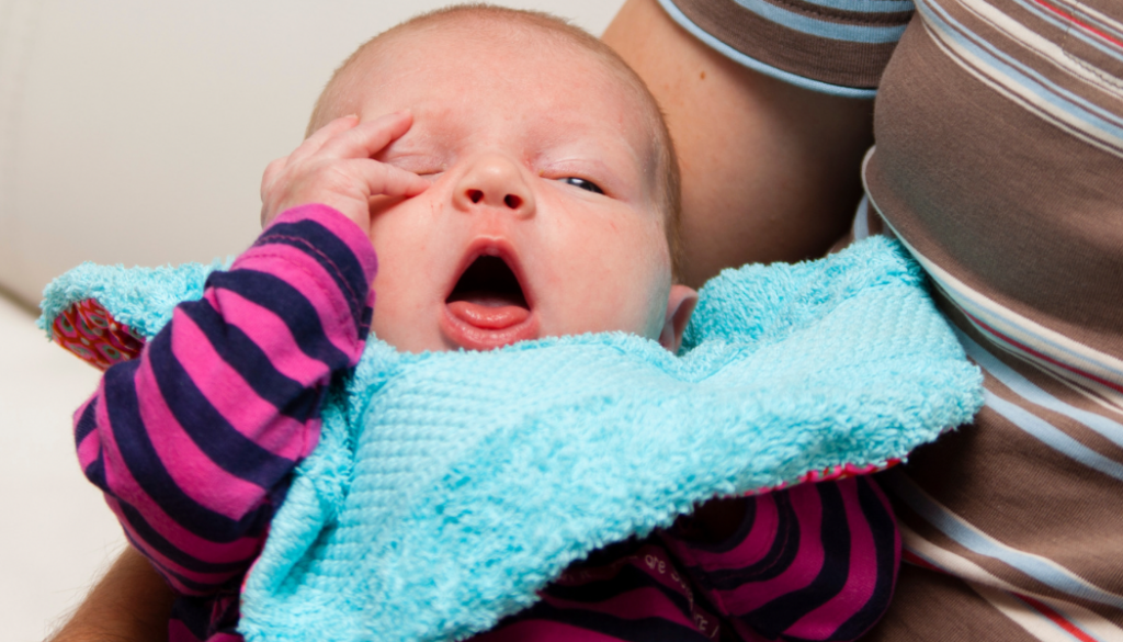 A newborn yawning and rubbing their eyes while being held, displaying common signs of overstimulation or tiredness during play. Recognizing infant cues like yawning, eye rubbing, and turning away helps parents respond before a baby becomes overwhelmed. Learning these signals is a key part of responsive newborn care.