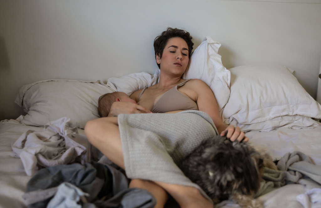 Exhausted postpartum mother breastfeeding newborn while lying in bed surrounded by unmade sheets, depicting the reality of postpartum overwhelm and baby blues