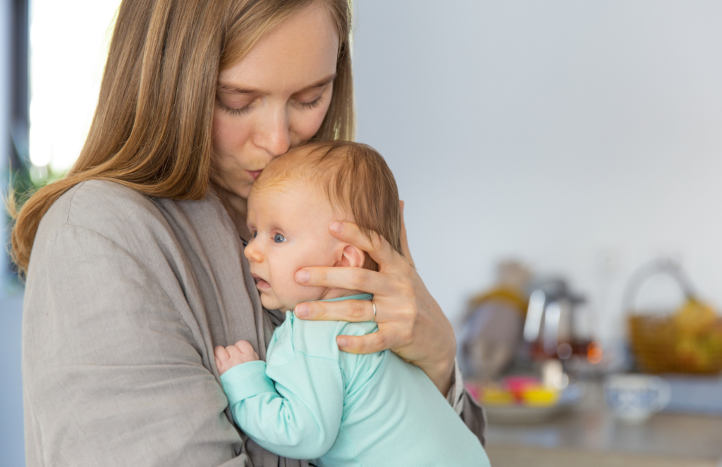 Mother gently kissing her alert baby on the top of the head at home, representing everyday bonding moments that matter despite the self-doubt that comes with mom guilt