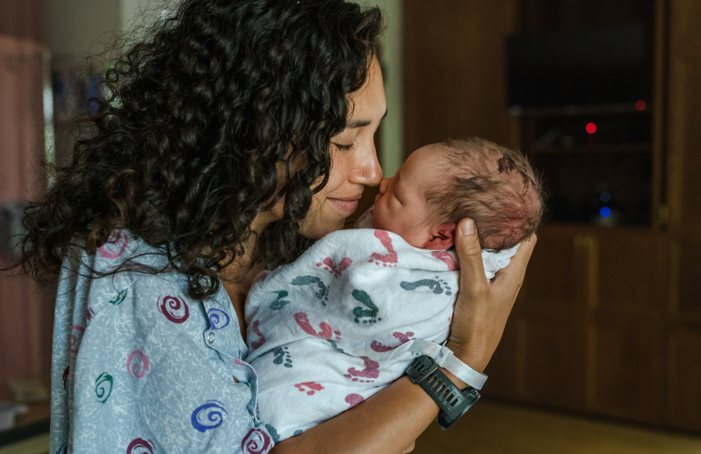 New mother in hospital gown nuzzling her swaddled newborn in the first moments after birth, representing the raw and tender beginning of the mother and baby bond