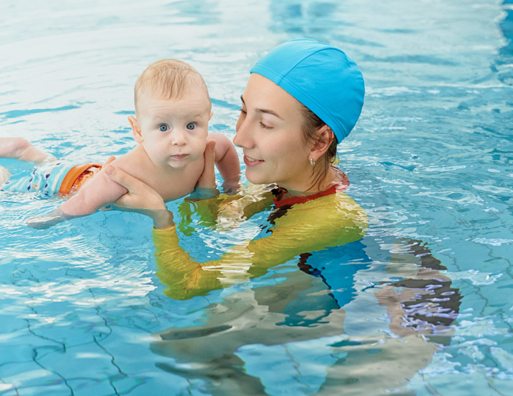 Photo of a smiling mother wearing a blue swim cap and yellow swimsuit holding her young baby in an indoor pool, with the baby in a striped orange swim diaper, demonstrating safe and supervised infant pool introduction.