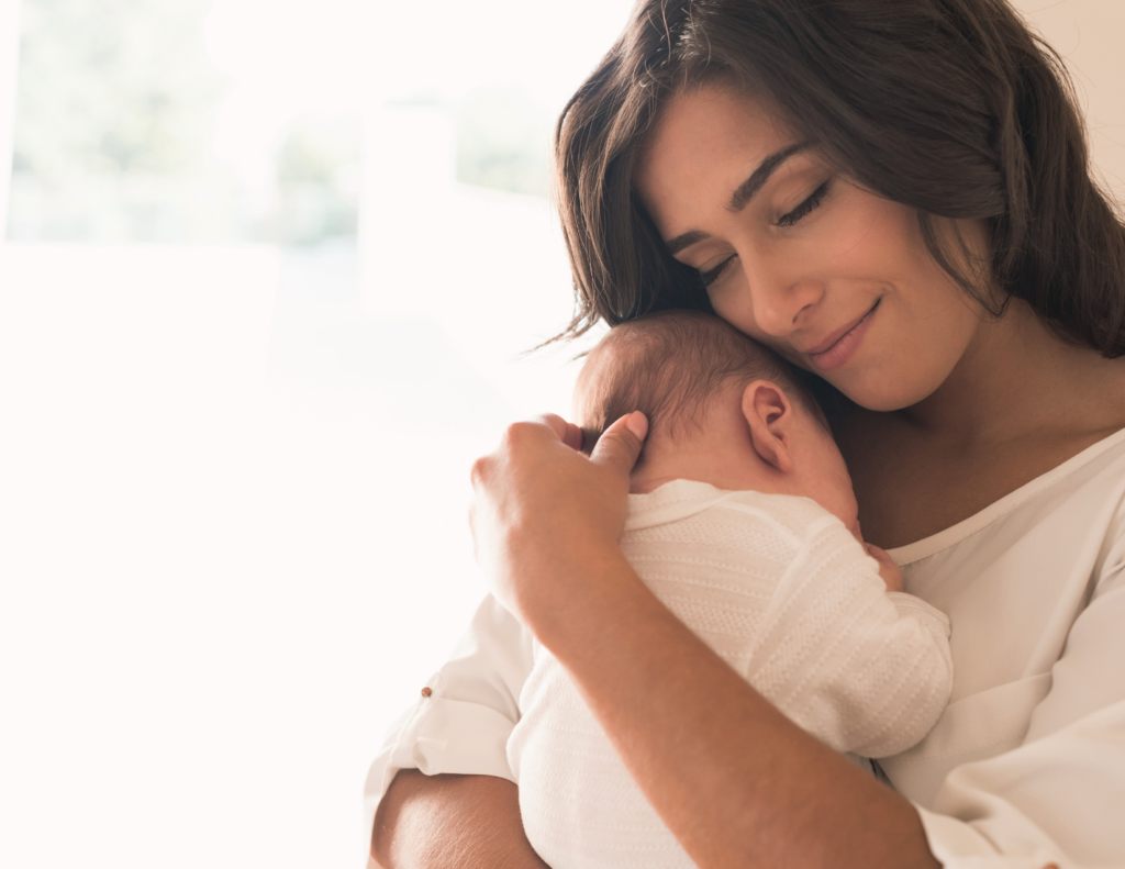 A smiling mother holding a sleeping newborn close to her cheek in a bright, softly lit room