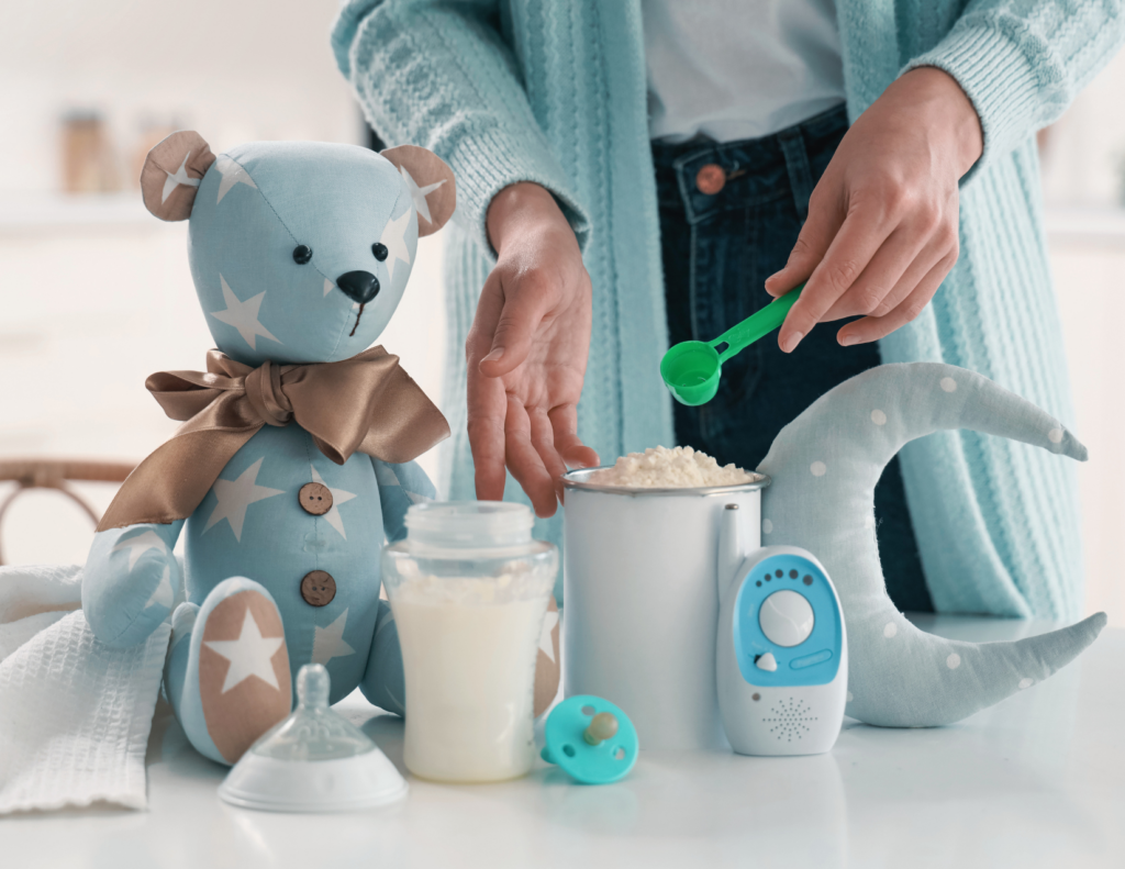 A parent scooping powdered infant formula into a bottle on a table with baby items including a teddy bear, pacifier, and baby monitor