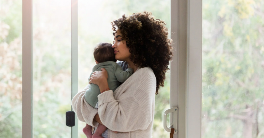 Postpartum mother standing by a bright window holding her baby close to her chest, representing the quiet emotional weight of mom guilt and self-doubt in early motherhood