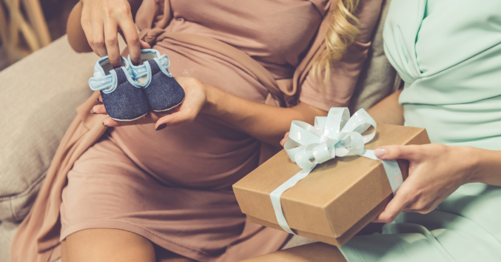 Pregnant woman holding tiny blue baby shoes while receiving a wrapped gift at a baby shower, representing traditional vs practical gift giving