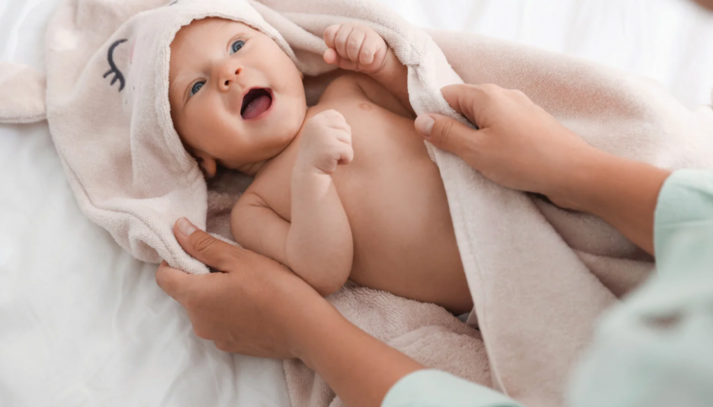  A smiling infant wrapped in a hooded towel being held by a caregiver after bath time. Responsive parenting moments like this strengthen the parent-infant bond and support healthy emotional development. Consistent, gentle interaction in early infancy lays the foundation for secure attachment and long-term emotional health.