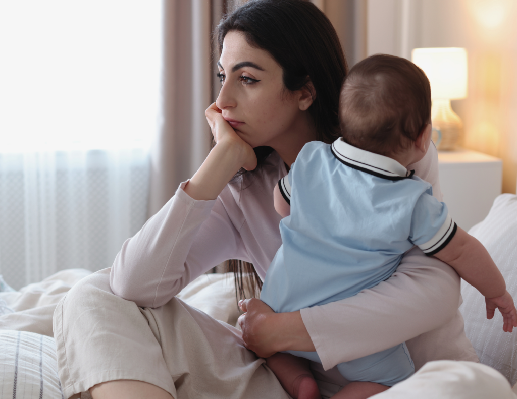 Young mother sitting on bed holding her baby while looking away with a tired, sad expression, representing postpartum depression and baby blues