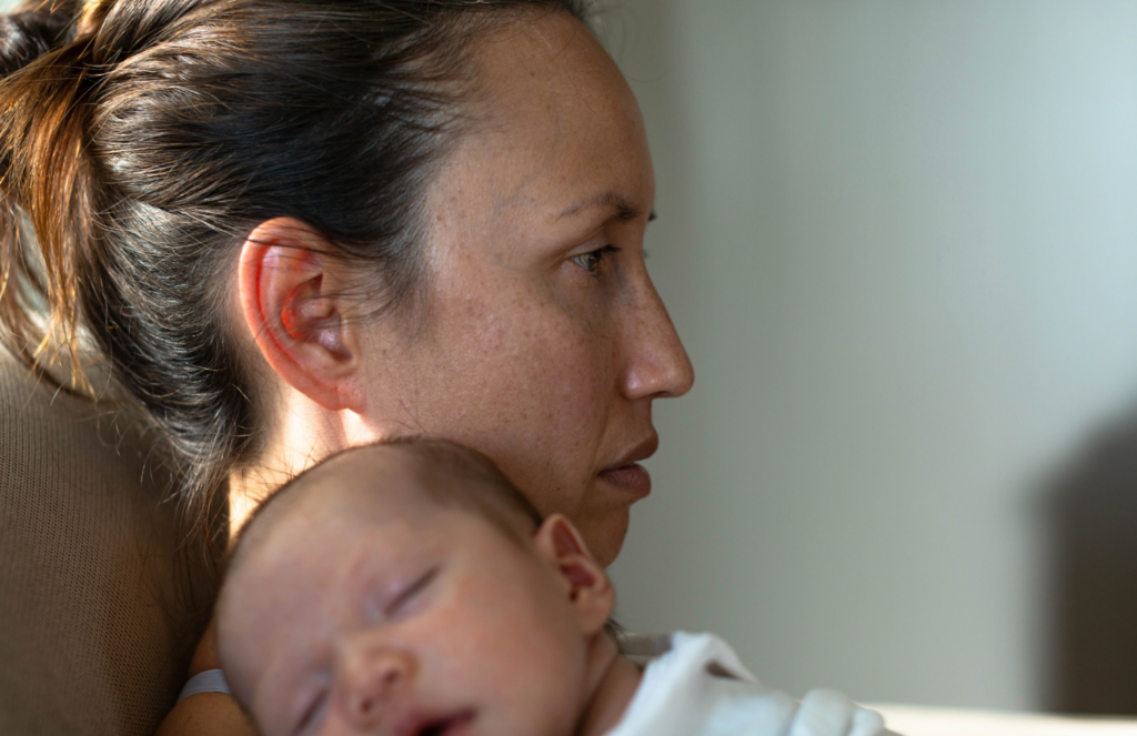 Tired postpartum mother holding sleeping newborn against her shoulder in profile, representing prolonged emotional exhaustion as a potential sign of postpartum depression