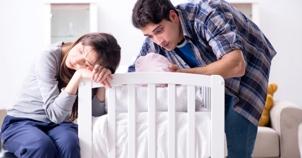  Sleep-deprived mother and father leaning over white crib together checking on their newborn baby 